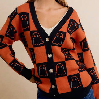 A woman wearing a vibrant orange and black checkered cardigan with ghost motifs, styled over a white top and blue jeans against a neutral beige background.