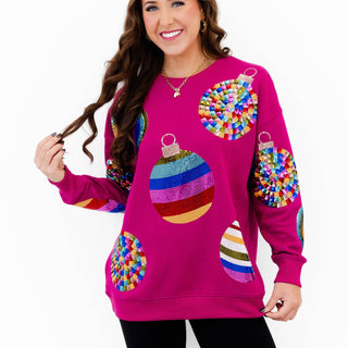 A woman modeling a bright magenta crewneck sweater decorated with colorful, textured Christmas ornament graphics, paired with black leggings, against a plain white background.