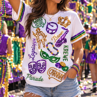 A woman wearing a white short-sleeve t-shirt with colorful Mardi Gras themed sequin graphics, including masks, a saxophone, a crown, and text like 'Mardi Gras' and 'Carnival', paired with denim shorts in a festive parade setting.