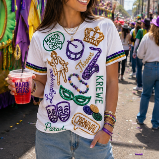 A woman wearing a white Mardi Gras graphic t-shirt with colorful sequined patches and lettering, standing on a parade street next to a decorated float.