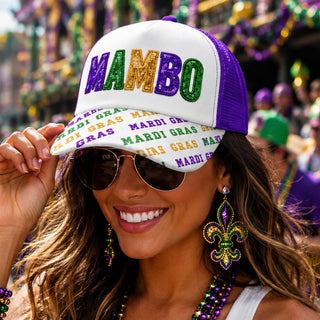 A woman wearing a white and purple trucker hat with 'MAMBO' in glittery Mardi Gras colors, along with sunglasses, beaded necklaces, and a fleur-de-lis earring, set against a festive Mardi Gras parade background.