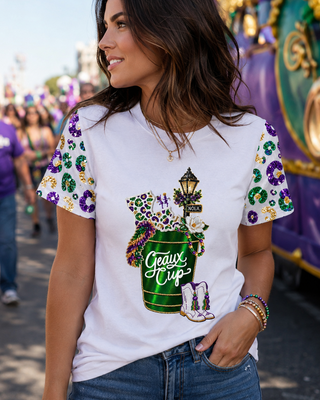 A woman wearing a white graphic t-shirt with Mardi Gras themed sleeve prints and a central 'Grave's Cup' bucket graphic, paired with blue jeans, standing outdoors in front of a festive parade float.