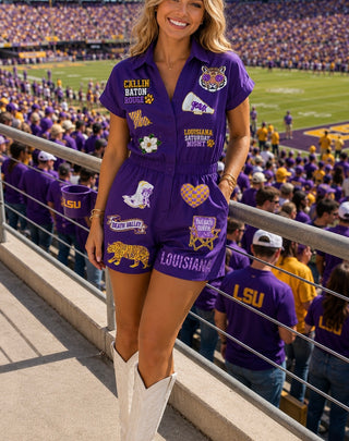 A woman stands in a stadium wearing a purple Louisiana Tigers themed short-sleeve romper with various team patches and white cowboy boots.