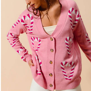 A woman wearing a pink knit cardigan with white and pink candy cane patterns, styled over a white top and white pants against a solid beige background.