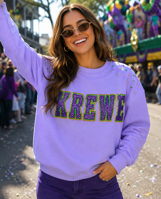 A woman wearing a lavender crewneck sweatshirt with 'KREWE' in purple and green Mardi Gras-style lettering, photographed outdoors at what appears to be a festival or parade.