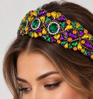 A close-up of a woman wearing an ornate Mardi Gras-style jeweled headband. The headband features large green, purple, and yellow gemstones with gold-tone metal settings and clear rhinestone accents, styled on her dark brown hair against a light gray background.