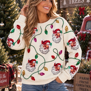 A woman wearing a festive Christmas crewneck sweater with sequined Santa faces and light bulbs, posed in front of a decorated Christmas tree and rustic holiday decorations.