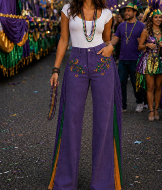A woman wearing purple wide-leg fringe pants with green and gold embroidered details and side fringe, styled with a white top and beaded accessories, photographed at a festive parade street event.