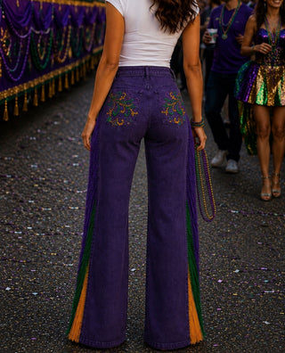 A person shown from the back wearing purple wide-leg jeans with colorful Mardi Gras fringe and embroidered details, standing on a street with parade decorations in the background.