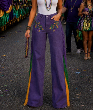 Purple wide-leg jeans with colorful Mardi Gras themed embroidery and green and gold fringe, worn by a model in a parade setting.