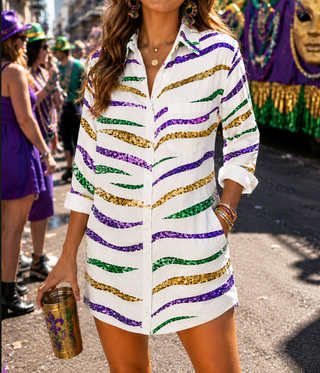 A woman wearing a white button-up shirt dress with wavy purple, green, and gold sequin stripes, standing outdoors at what appears to be a Mardi Gras parade. She is holding a gold tumbler with purple and green graphics.