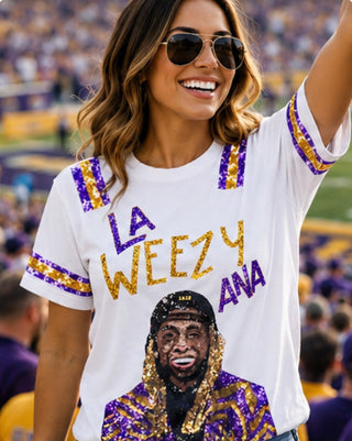 A woman wearing a white graphic T-shirt with purple and gold sequin-style lettering that reads 'LA WEEZY ANA' and a printed portrait graphic on the front, photographed in a crowded stadium setting.